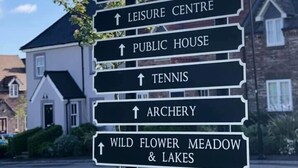 Un panneau de signalisation indiquant différentes directions à l’aide de multiples flèches, pointant vers divers lieux tels que la réception, le service des ventes, le terrain d’entraînement au golf, la plage, le centre de loisirs, un pub, les courts de tennis, le terrain d’archerie, ainsi que la prairie fleurie et les lacs.