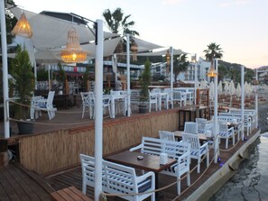 Un área de comedor al aire libre con sillas y mesas blancas, sombrillas y luces colgantes.