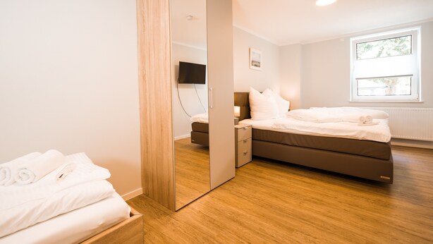 Une chambre moderne avec un plancher en bois, un lit recouvert de draps blancs, une table de chevet, un miroir et une fenêtre laissant entrer la lumière du jour.