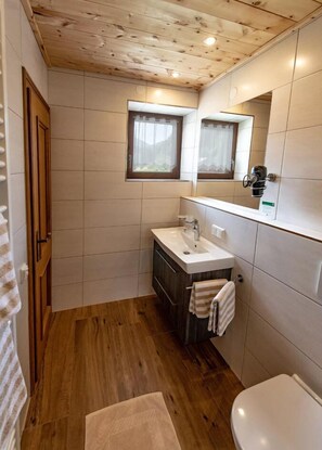 A bathroom with a wooden ceiling, white tiles, a wooden vanity, a white sink, a mirror, and a toilet. - Hinterkellaubauer (Kuchl)