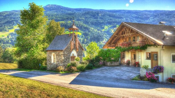 Ländliche Szenerie mit einer Steinkirche, einem Holzhaus mit Satteldach und freiem Blick auf die umliegenden Berge.