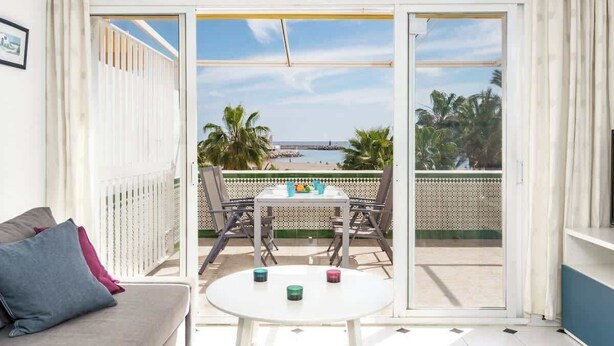 Un balcón con mesa y sillas, un sofá y vistas a la playa y palmeras.