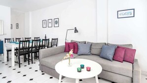 Un salón moderno con un sofá gris, una mesa de centro redonda blanca y una zona de comedor con una mesa blanca y sillas negras.