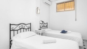 Una habitación con dos camas, una mesita, aire acondicionado instalado en la pared y una ventana con persianas.