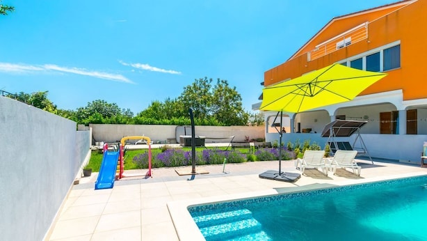 A swimming pool with a slide and lounge chairs, surrounded by a garden and a building with a yellow awning. - Apartment Janko (Medulin)