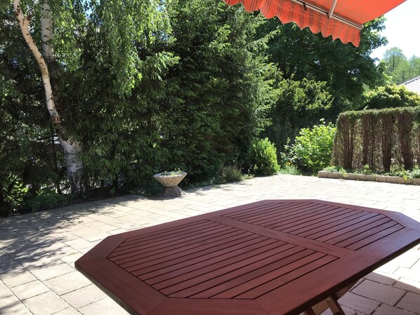 Un espace patio avec une table en bois, un bac à fleurs en pierre et un parasol rouge.