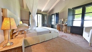 Un dormitorio con una cama grande, una lámpara de noche, una mesita de mimbre y vistas al verdor por la ventana.