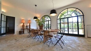 Un comedor con una mesa grande, varias sillas y lámparas colgantes. Hay ventanas amplias con arcos y vistas a espacios verdes al exterior.
