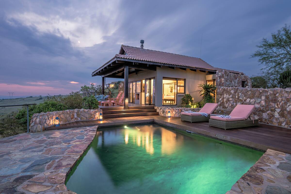 A poolside area with a stone wall, lounge chairs, and a building with a tiled roof.