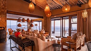 A spacious living area with a large sofa, a coffee table, and a wicker chair. There are decorative vases on a shelf and a view of the outdoors through large windows.