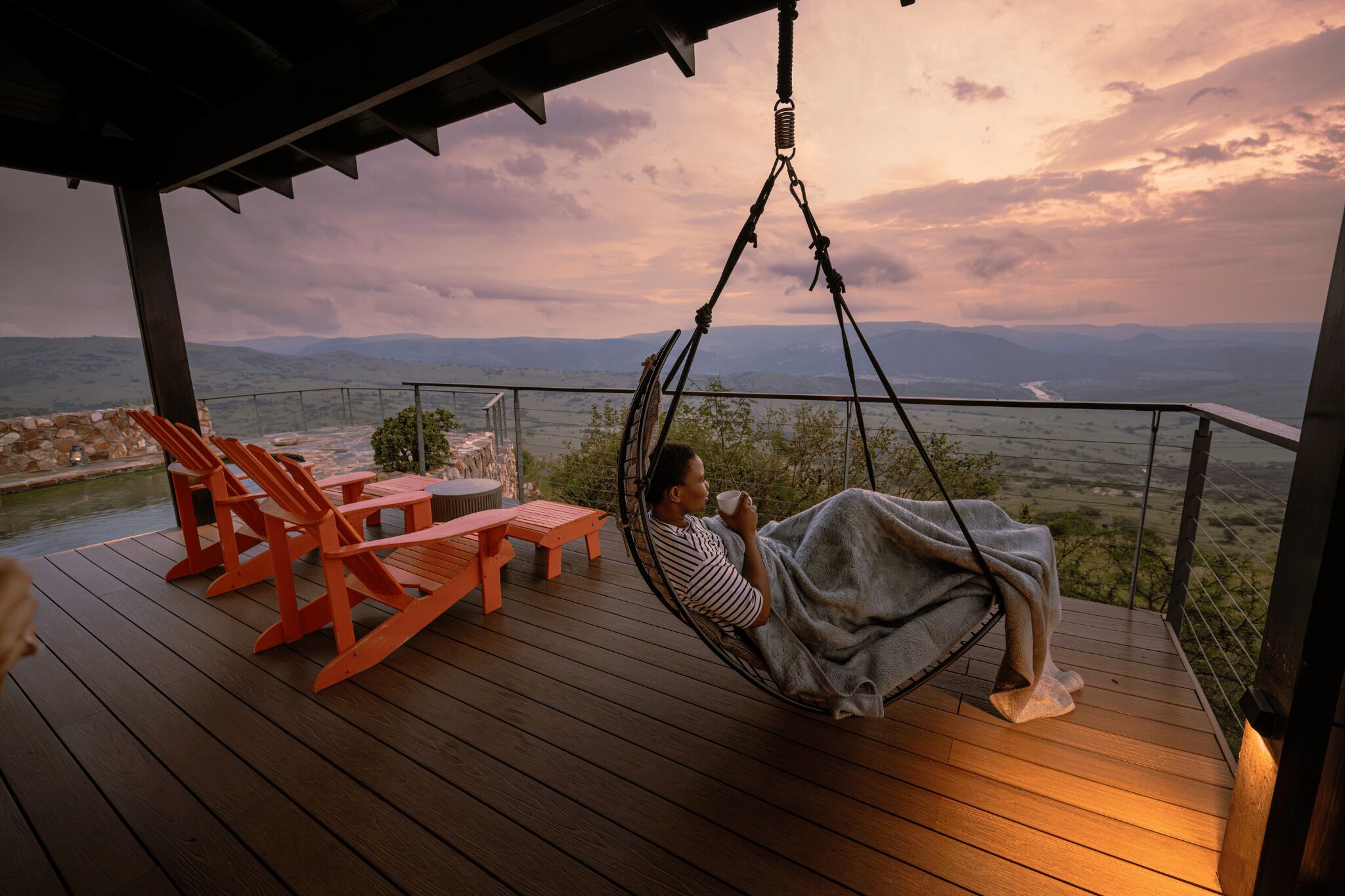 Une personne se détend dans un fauteuil suspendu sur une terrasse en bois, avec vue sur le paysage.