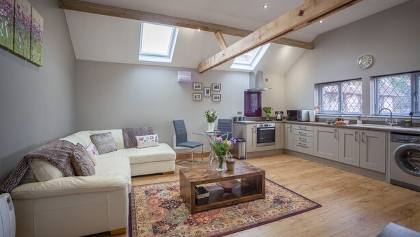 A modern living room with a sofa, coffee table, and a kitchen area with a washing machine. - HEREFORDSHIRE – Little Oak: Charming Rural Cottage, Sleeps 2, Cozy Getaway (HEREFORD)