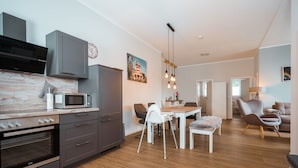 A modern kitchen with built-in appliances, a dining area with a white table and chairs, and a living room with a sofa and armchairs. - Haus Seeweg Geierswalde - Apartment 2 (Elsterheide)