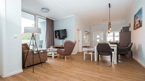A modern living room with a sofa, a dining table with chairs, a TV mounted on the wall, and a kitchen area with a refrigerator and sink. - Haus Seeweg Geierswalde - Apartment 2 (Elsterheide)