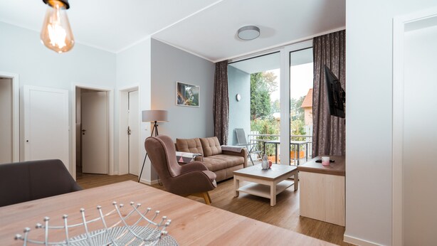 A modern living room with a wooden dining table, a sofa, and a coffee table. There is a TV mounted on the wall and a window with curtains. - Haus Seeweg Geierswalde - Apartment 2 (Elsterheide)