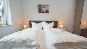 Una cama doble con sábanas blancas, dos mesitas de noche con lámparas, un cuadro enmarcado en la pared y una ventana con cortinas.