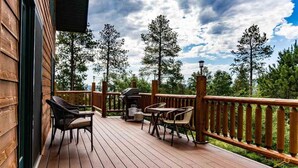 Una terraza de madera con mobiliario exterior, una parrilla y vistas a árboles y al cielo.