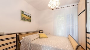 A bedroom with a bed, a nightstand, a chair, a window with curtains, and a painting on the wall. - Beautiful view apartment Loreta (Labin)