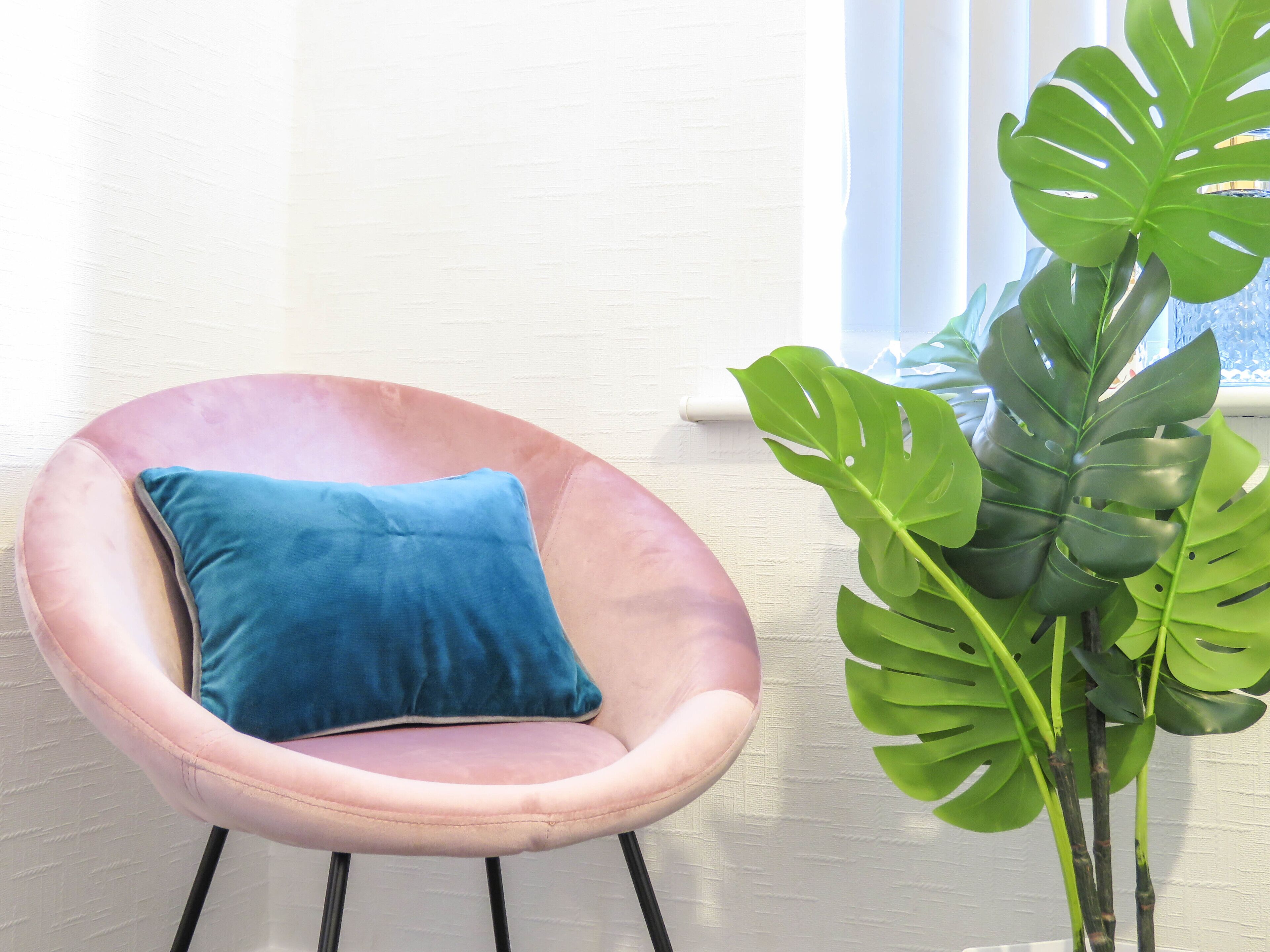 Una poltrona rosa con un cuscino blu e una grande pianta di monstera verde accanto.