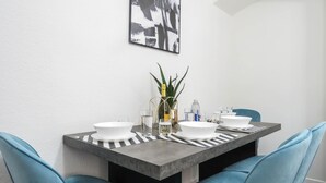 Een eetgedeelte met een rechthoekige tafel, twee blauwe stoelen, een vaas met een plant en een schilderij aan de muur.
