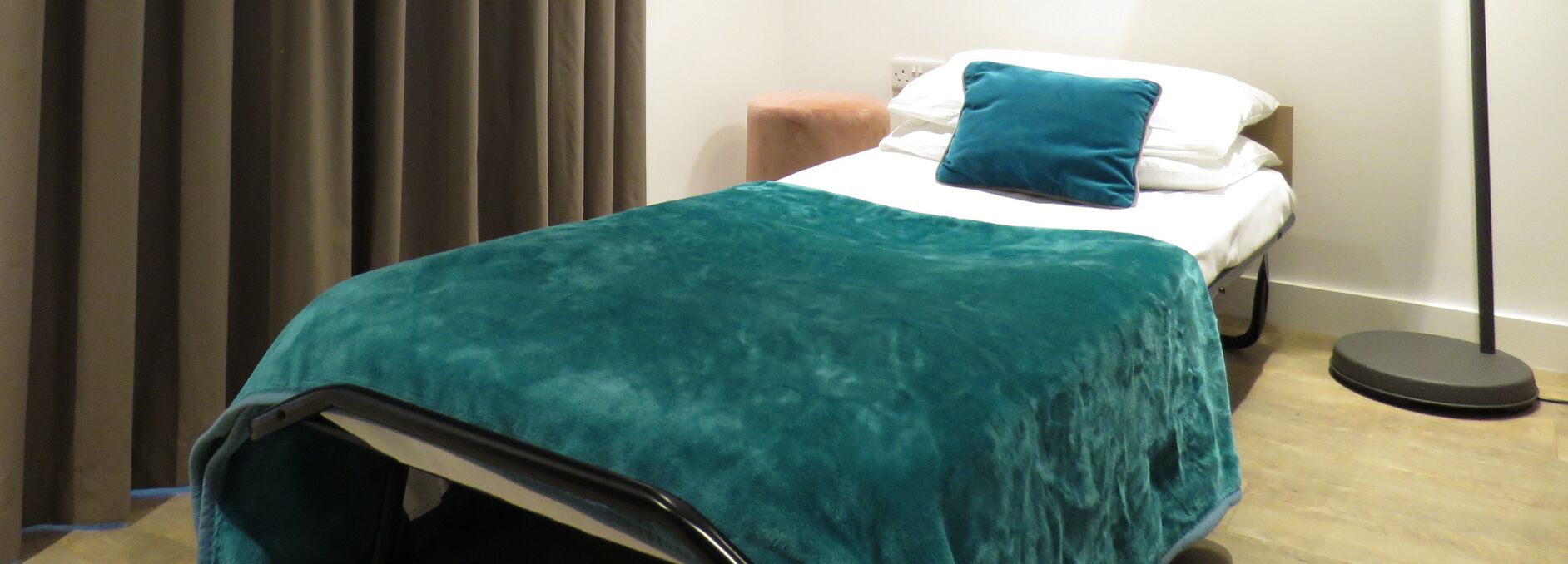 A single bed with a teal blanket, a white pillow, and a blue pillow.