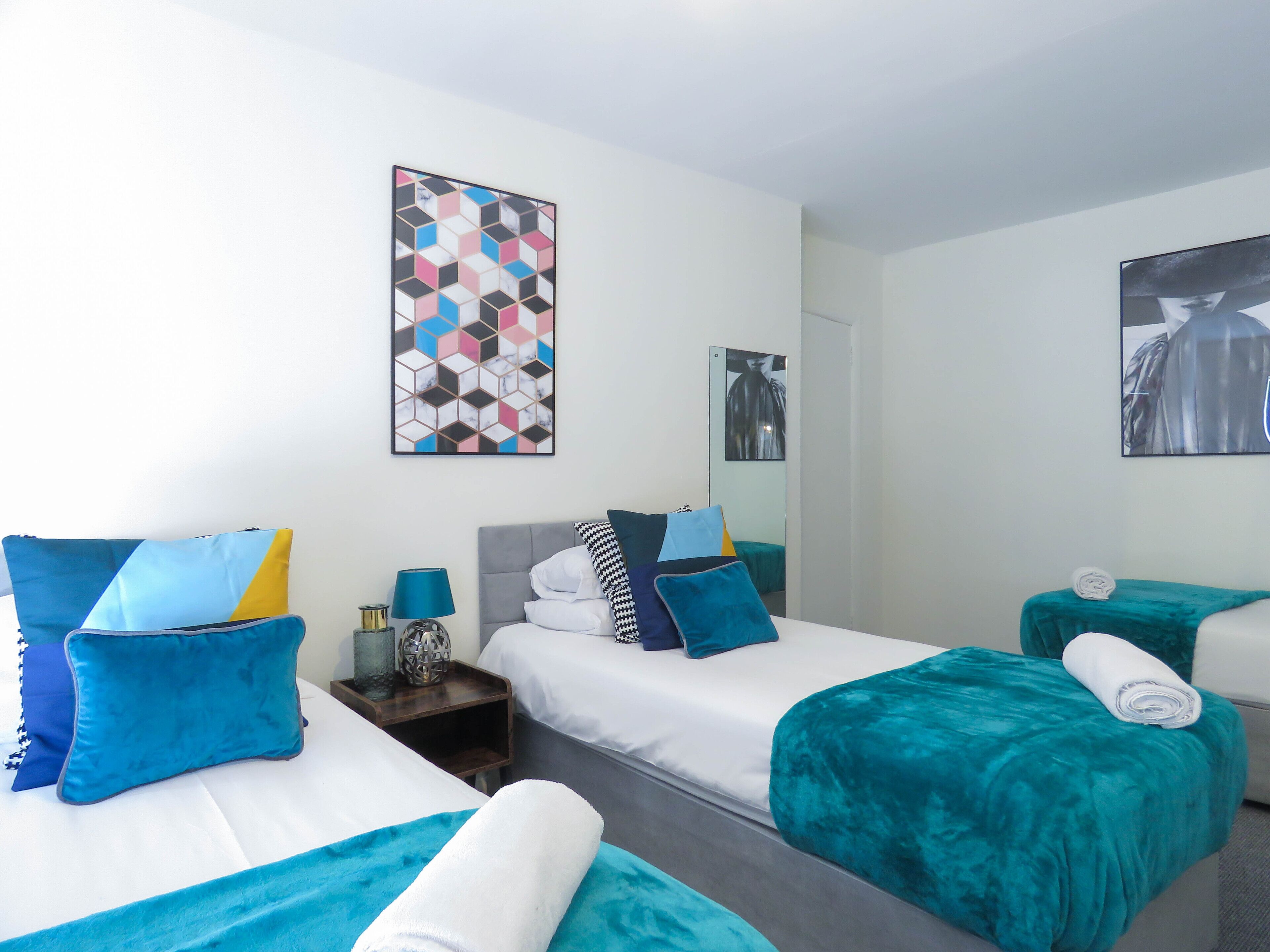 A hotel room with two beds, each with teal and white bedding, a large abstract painting, and a mirror reflecting a person.
