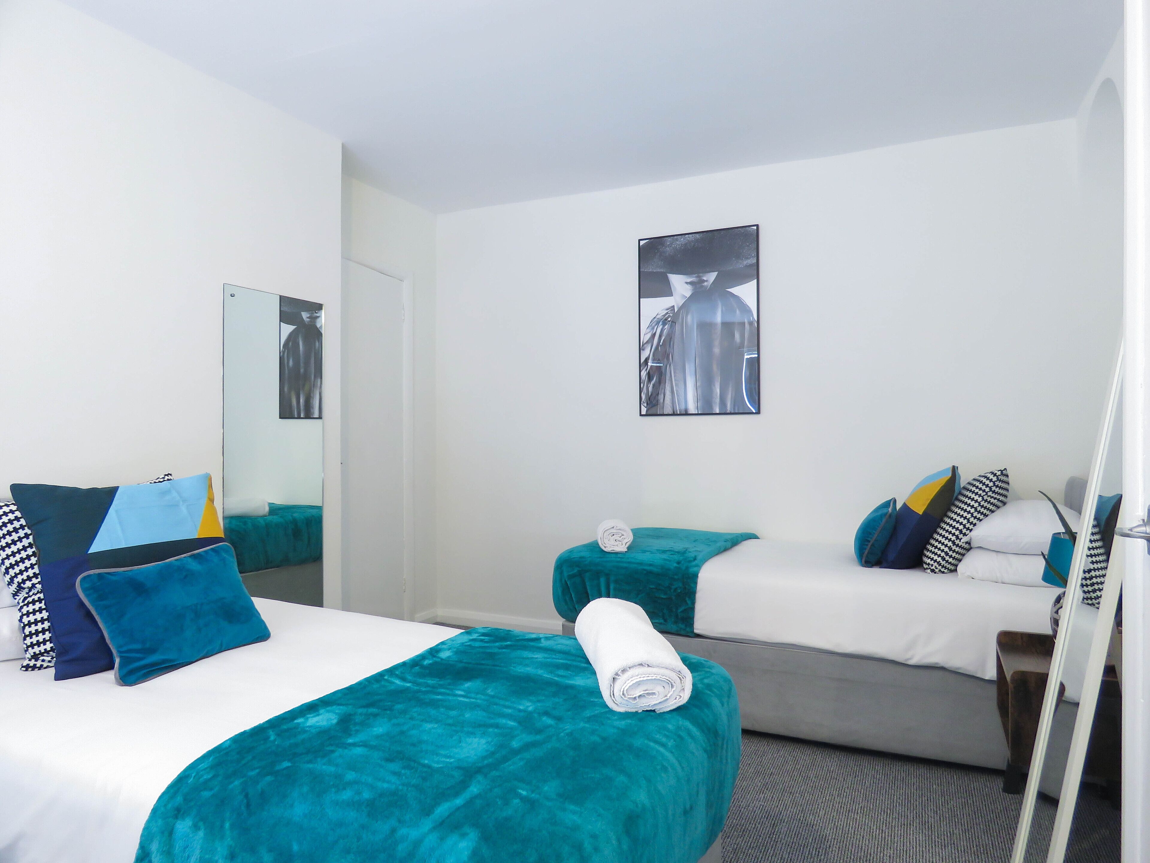Ein Hotelzimmer mit zwei Betten, jeweils mit türkisfarbenem Bettzeug und Kissen, einem großen Spiegel und einem gerahmten Kunstwerk an der Wand.
