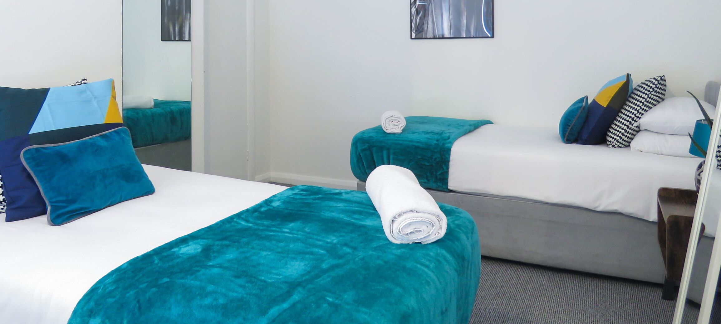 A hotel room with two beds, each with teal bedding and multiple pillows. There is a mirror on the wall and a small table with a towel.