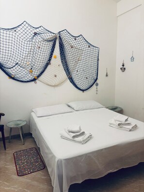 A neatly made bed with white linens, a small round table, and a decorative wall hanging.
