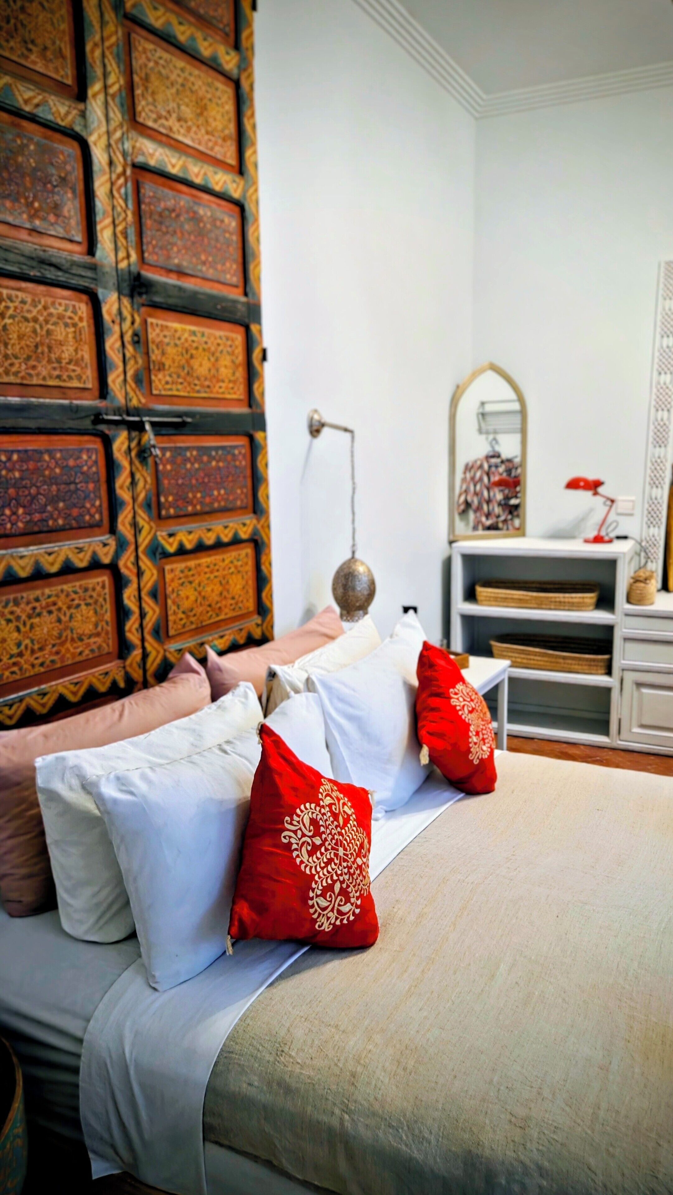 Una cama bien hecha con almohadas blancas y rojas, un cabecero estampado y una mesita de noche con una lámpara.