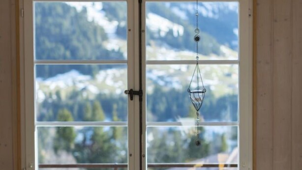 A room with a wooden interior, a white table, and four chairs. There are two windows with a view of mountains and trees. - Guesthouse Alive (Adelboden)