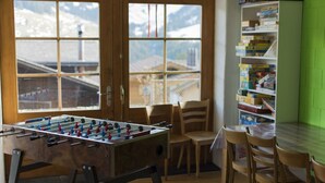 A room with a foosball table, a bookshelf, and a view of a snowy mountain. - Guesthouse Alive (Adelboden)