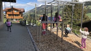 Children playing on a playground with a metal structure, surrounded by a paved area and a house in the background. - Guesthouse Alive (Adelboden)