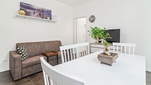 A modern living room with a sofa, dining table, chairs, a potted plant, a flat-screen TV, and a painting on the wall.