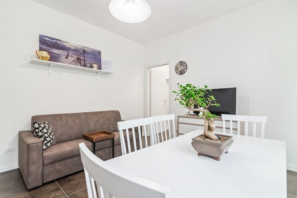 A modern living room with a sofa, dining table, chairs, a potted plant, a flat-screen TV, and a painting on the wall.