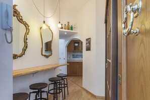 Un couloir avec une porte en bois, un miroir, un luminaire et un petit coin cuisine doté de meubles en bois et d’une étagère.