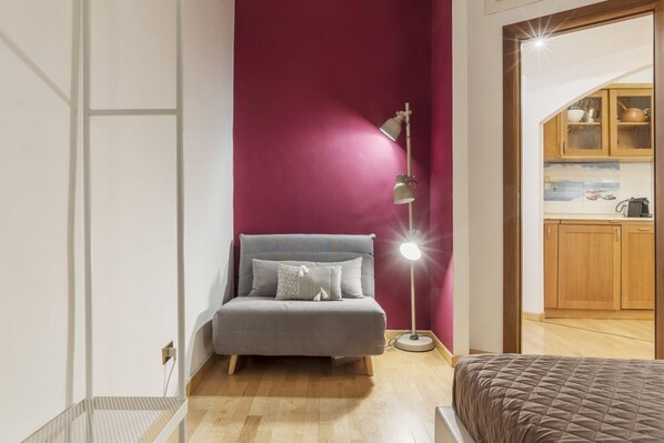 Une pièce avec un canapé gris, un lampadaire, un meuble en bois et une vue sur une cuisine.