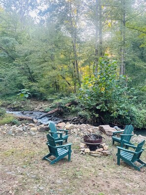 A natural outdoor setting with a small stream, surrounded by dense foliage and trees. There are three green Adirondack chairs arranged around a fire pit.