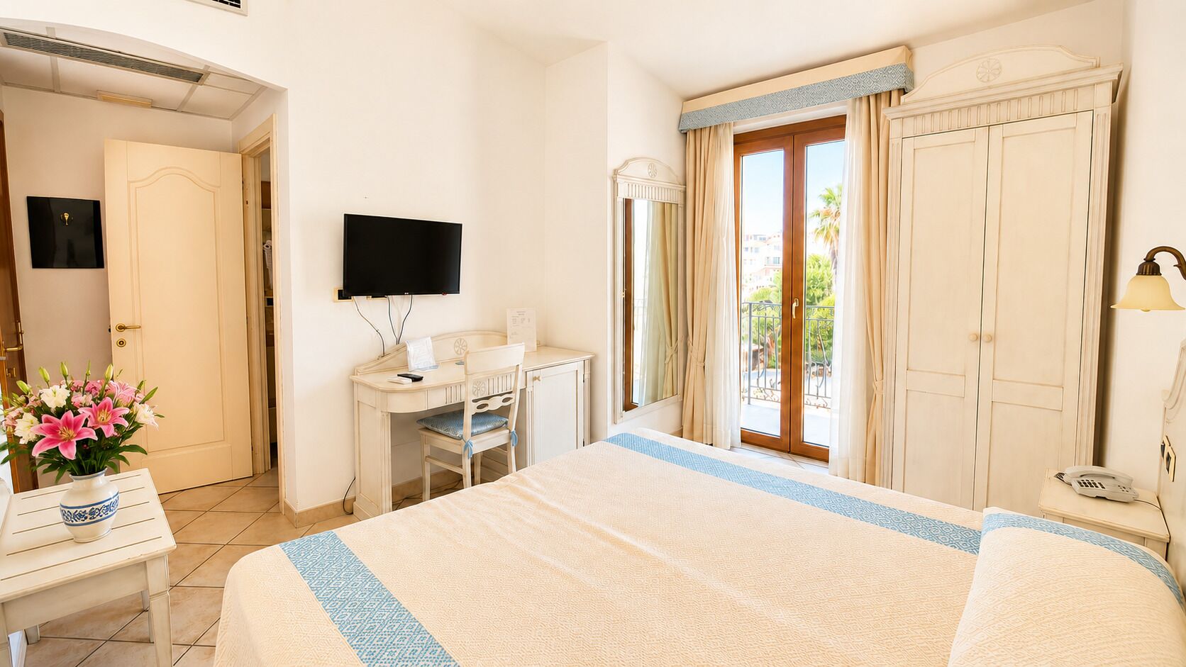 A hotel room with a bed, a desk, a television, a window with curtains, and a vase of flowers.