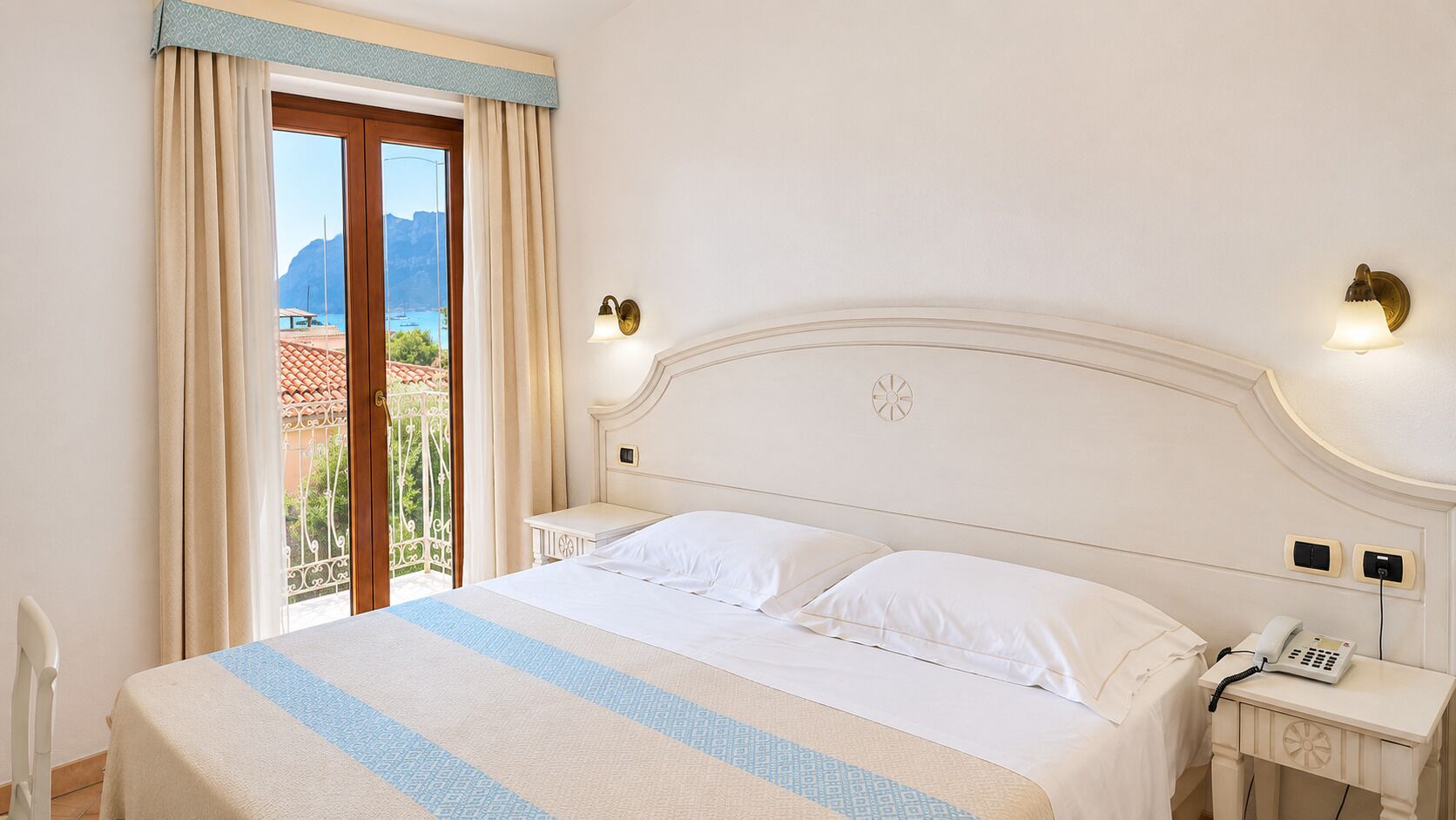 A hotel room with a bed, a nightstand with a phone, and a view of the outdoors.