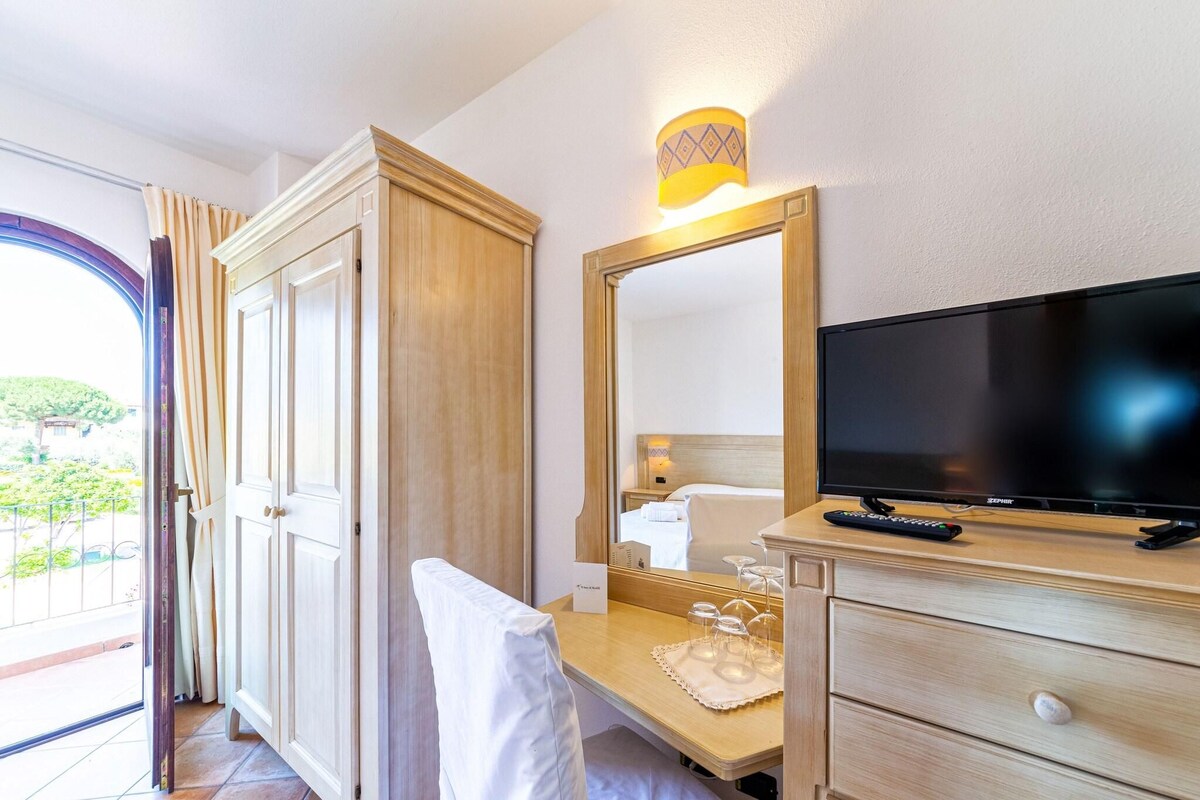 A hotel room with a wooden wardrobe, a bedside table with a television, a mirror, and a window with curtains.