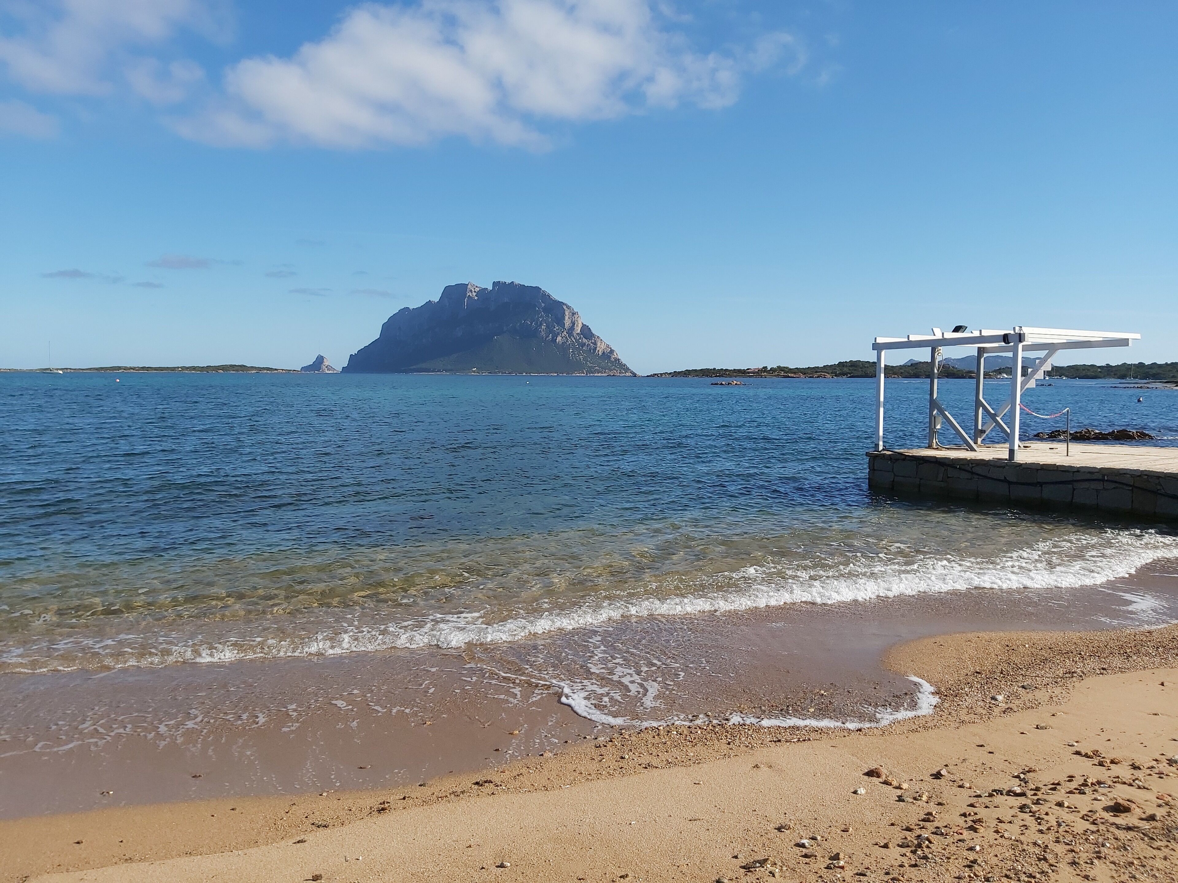 Une plage à l’eau claire, un promontoire rocheux à l’horizon et une structure blanche sur la droite.