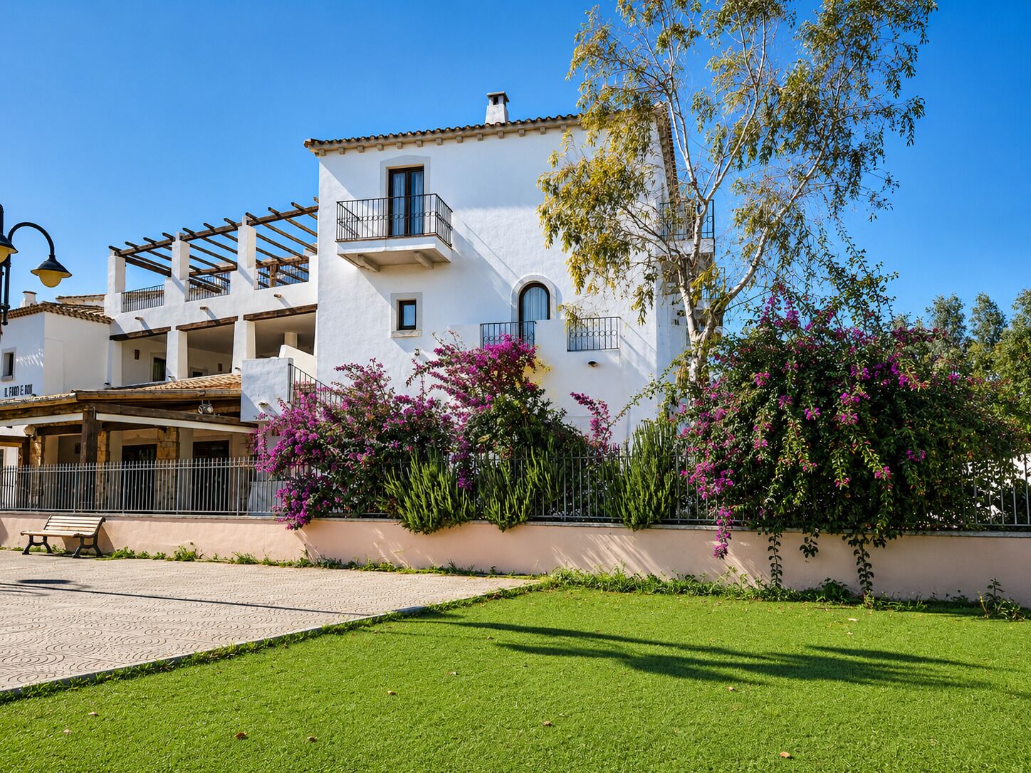 A white building with balconies, a paved walkway, and a grassy area with pink flowers.