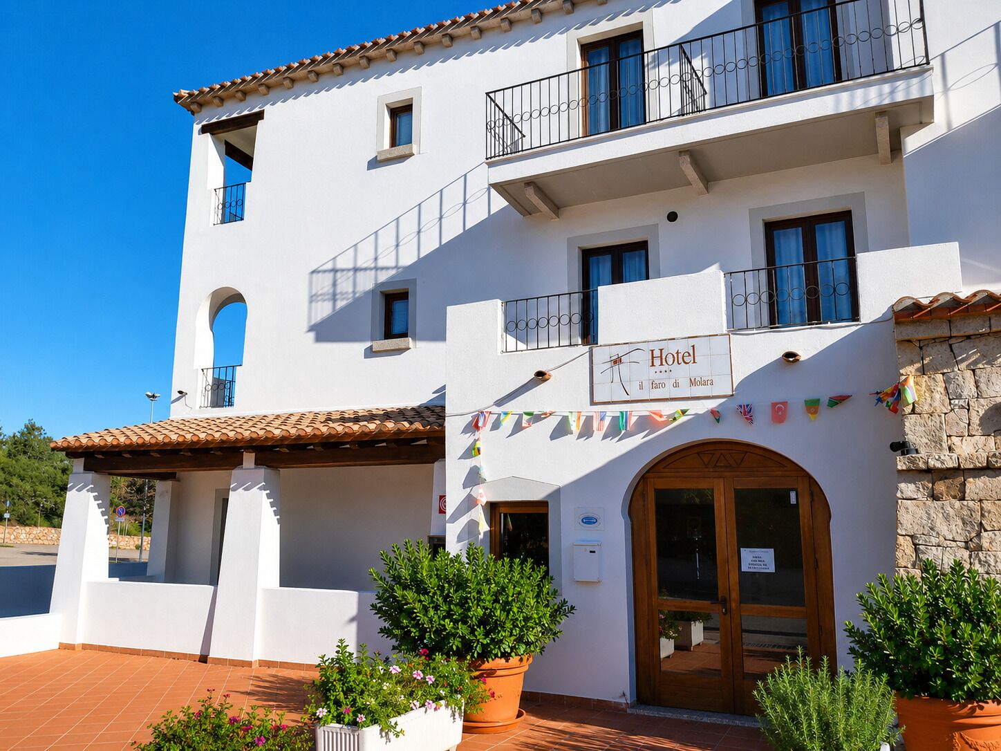 A white building with a sign indicating it's a hotel, featuring a tiled roof, balconies, and potted plants.
