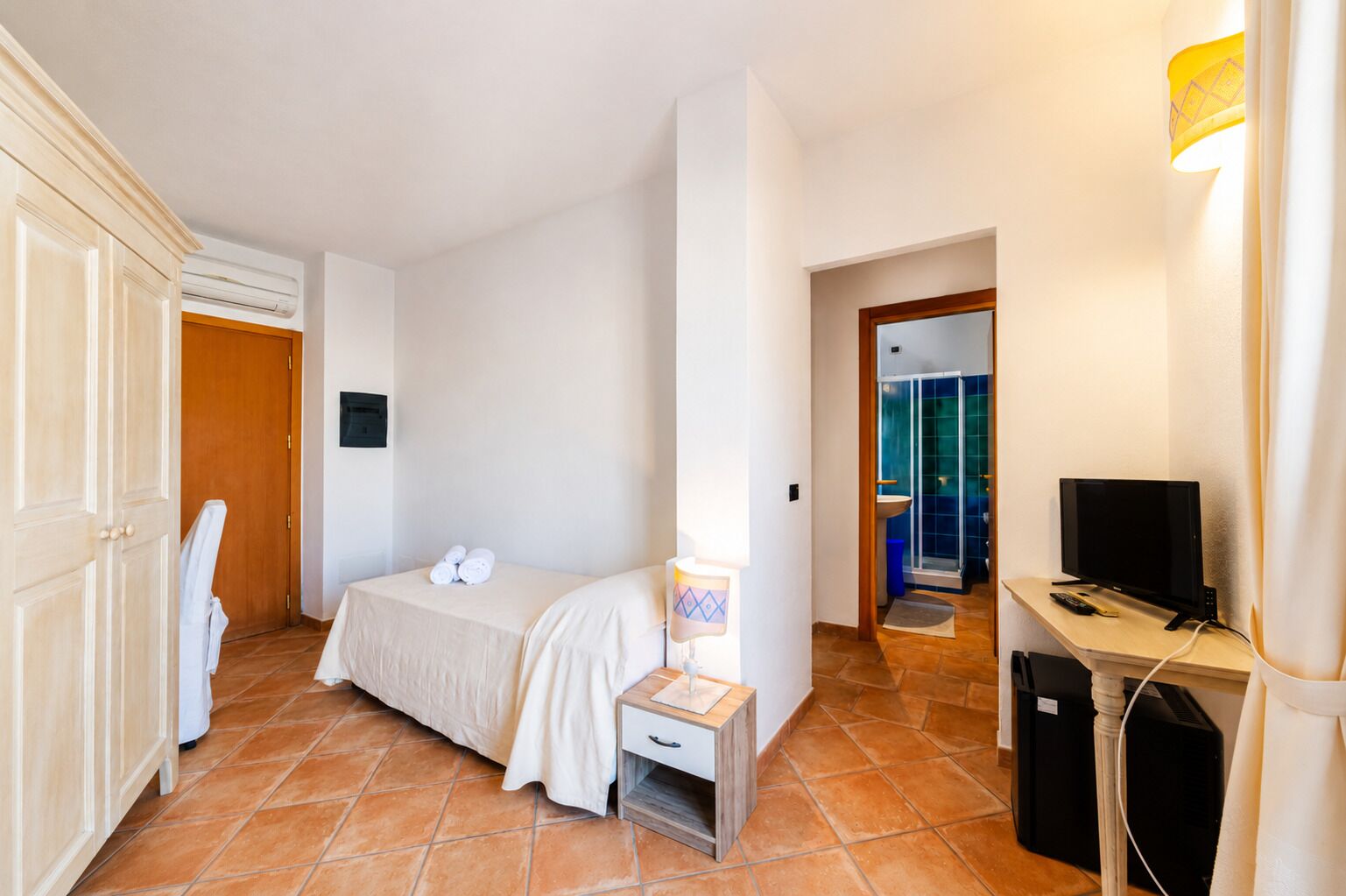 Habitación de hotel con cama, mesita de noche con lámpara, televisor y zona de ducha.