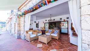 A covered outdoor seating area with wicker furniture, a stone wall, and a view of the street. - Hotel Il Faro di Molara (Loiri Porto San Paolo)