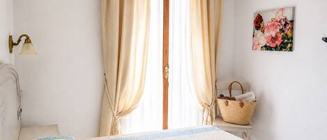 Un dormitorio con cama, cortinas, lámpara, un cuadro y una cesta.