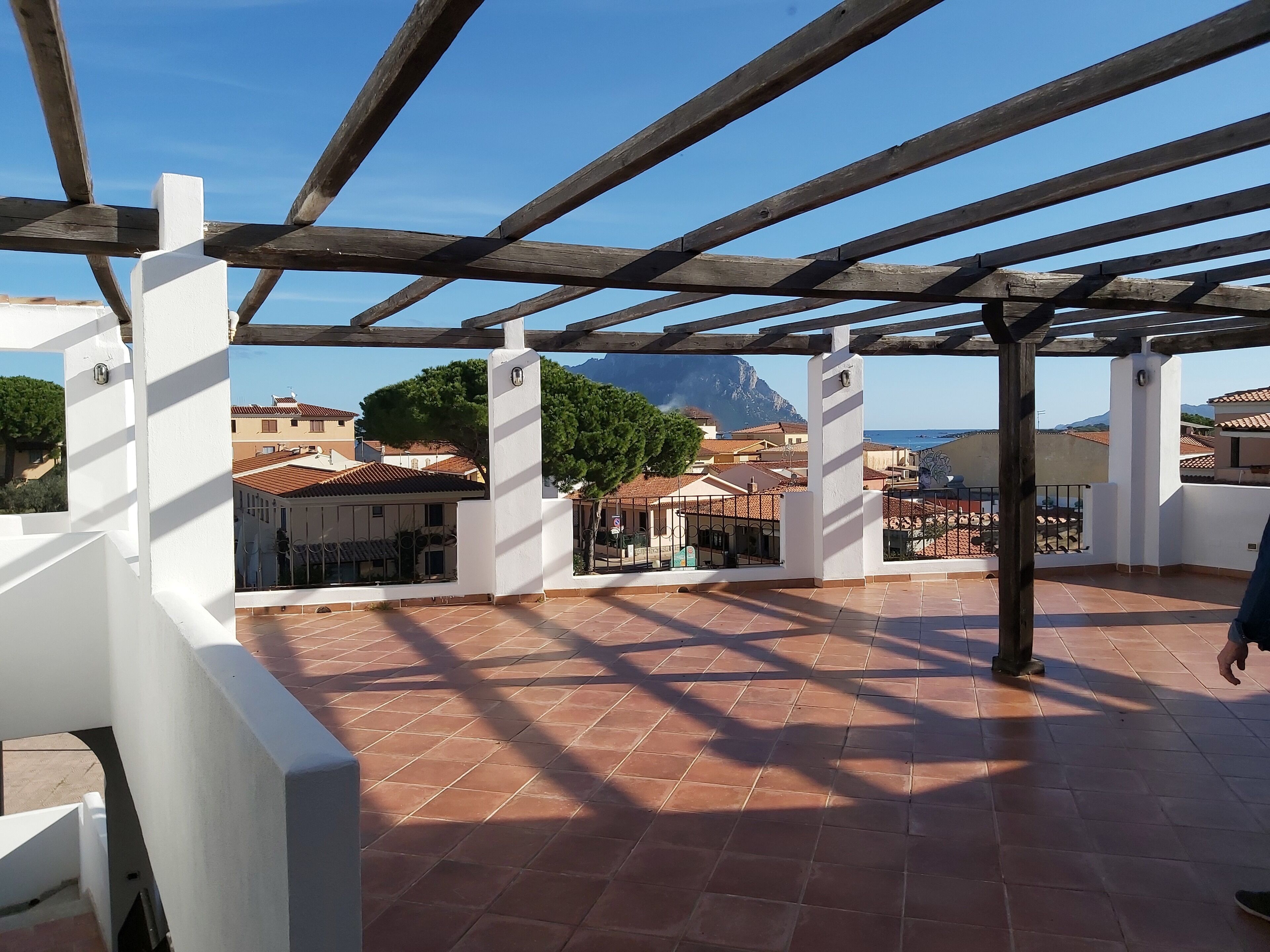 Una terraza al aire libre techada con suelo de baldosas, vigas de madera y pilares blancos.