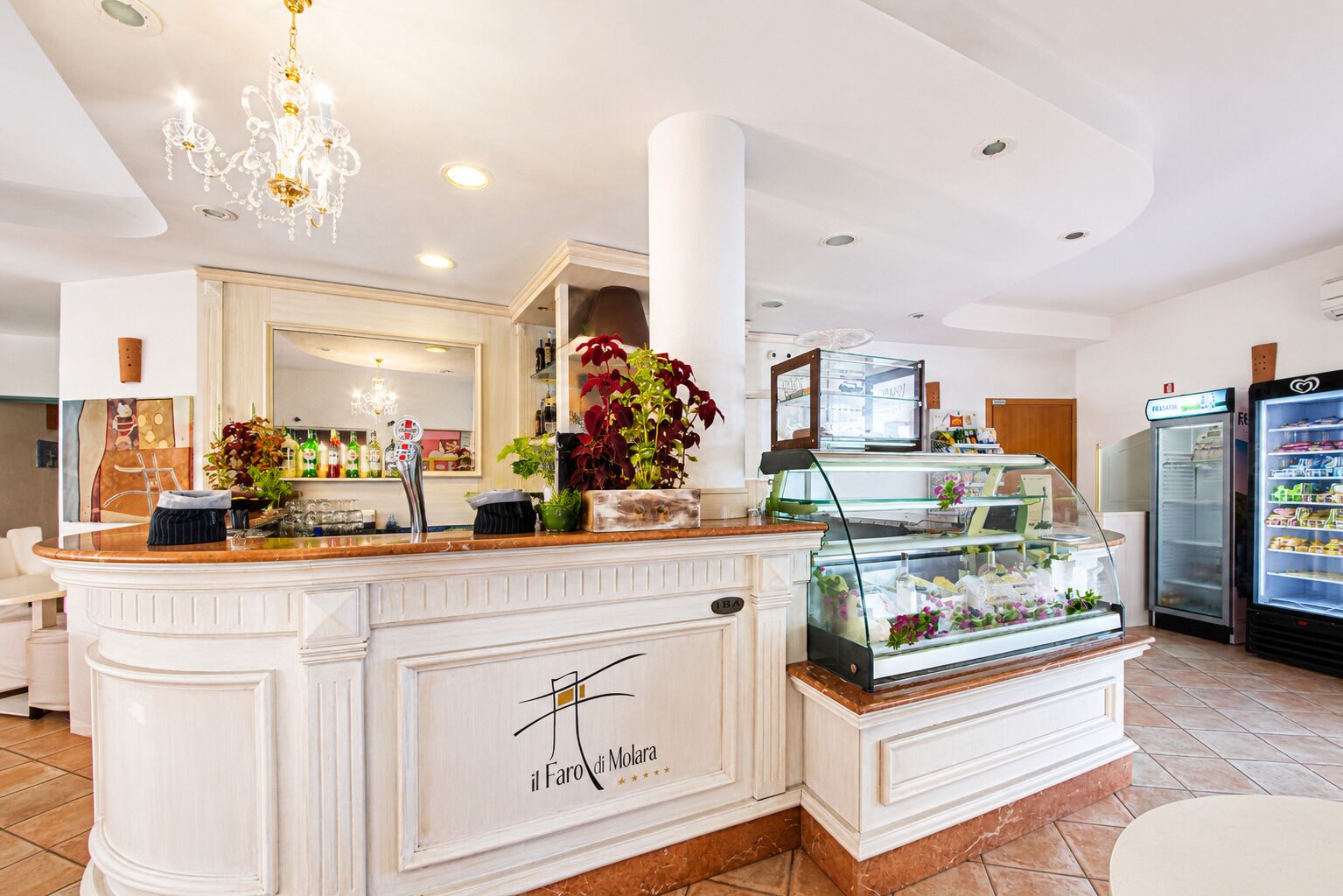 Un vestíbulo de hotel con mostrador de recepción, un candelabro, un refrigerador y una vitrina con flores.
