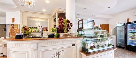 Un vestíbulo de hotel con mostrador de recepción, un candelabro, un refrigerador y una vitrina con flores.